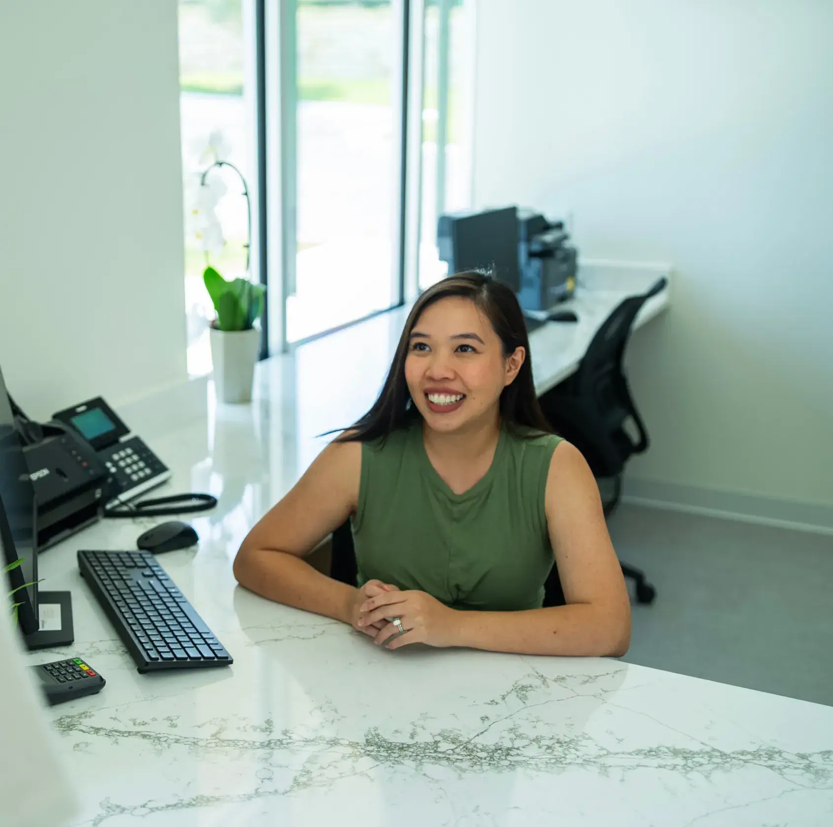 A woman in a green shirt sits smiling at a white reception desk with a computer.