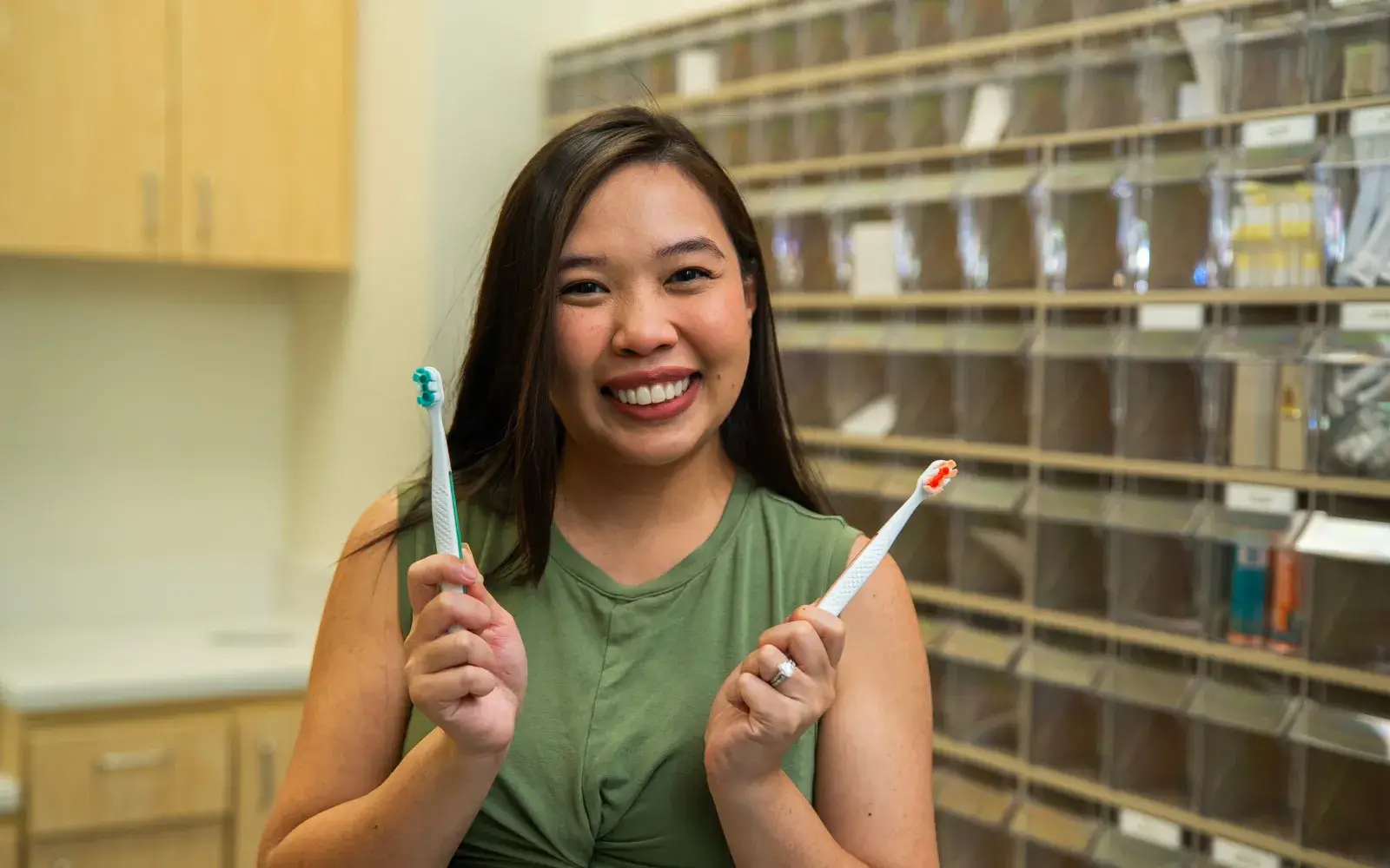 A woman smiling and holding two toothbrushes in a room with shelves behind her.