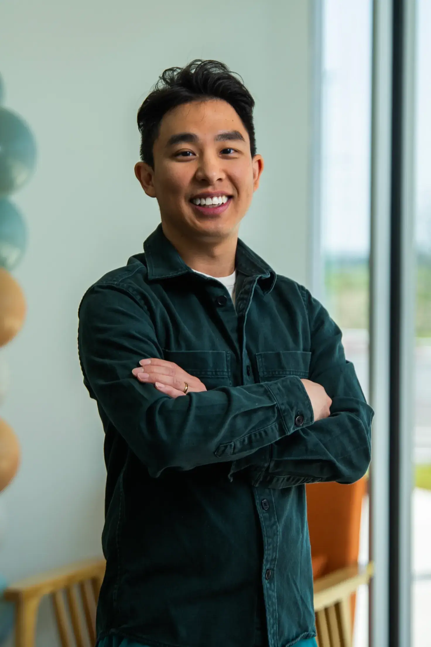 A person stands smiling and looking forward with arms crossed by a window.