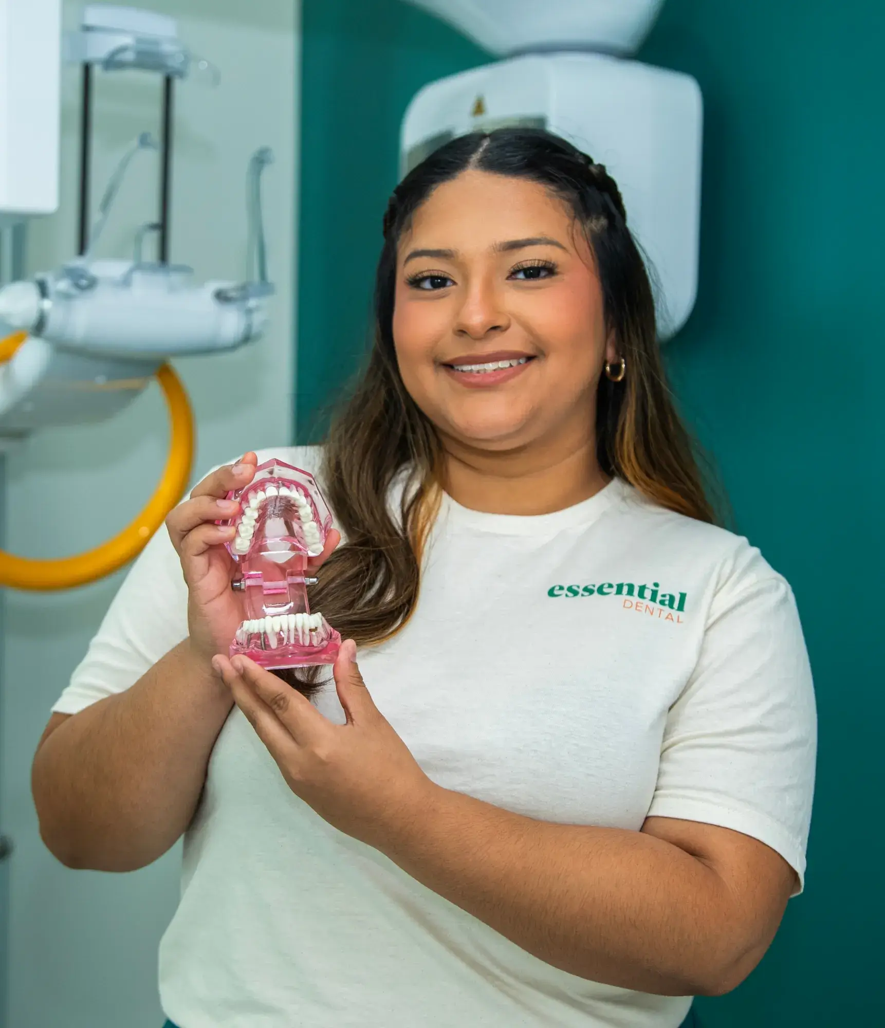 A smiling woman holds a dental model, standing in a dental office.