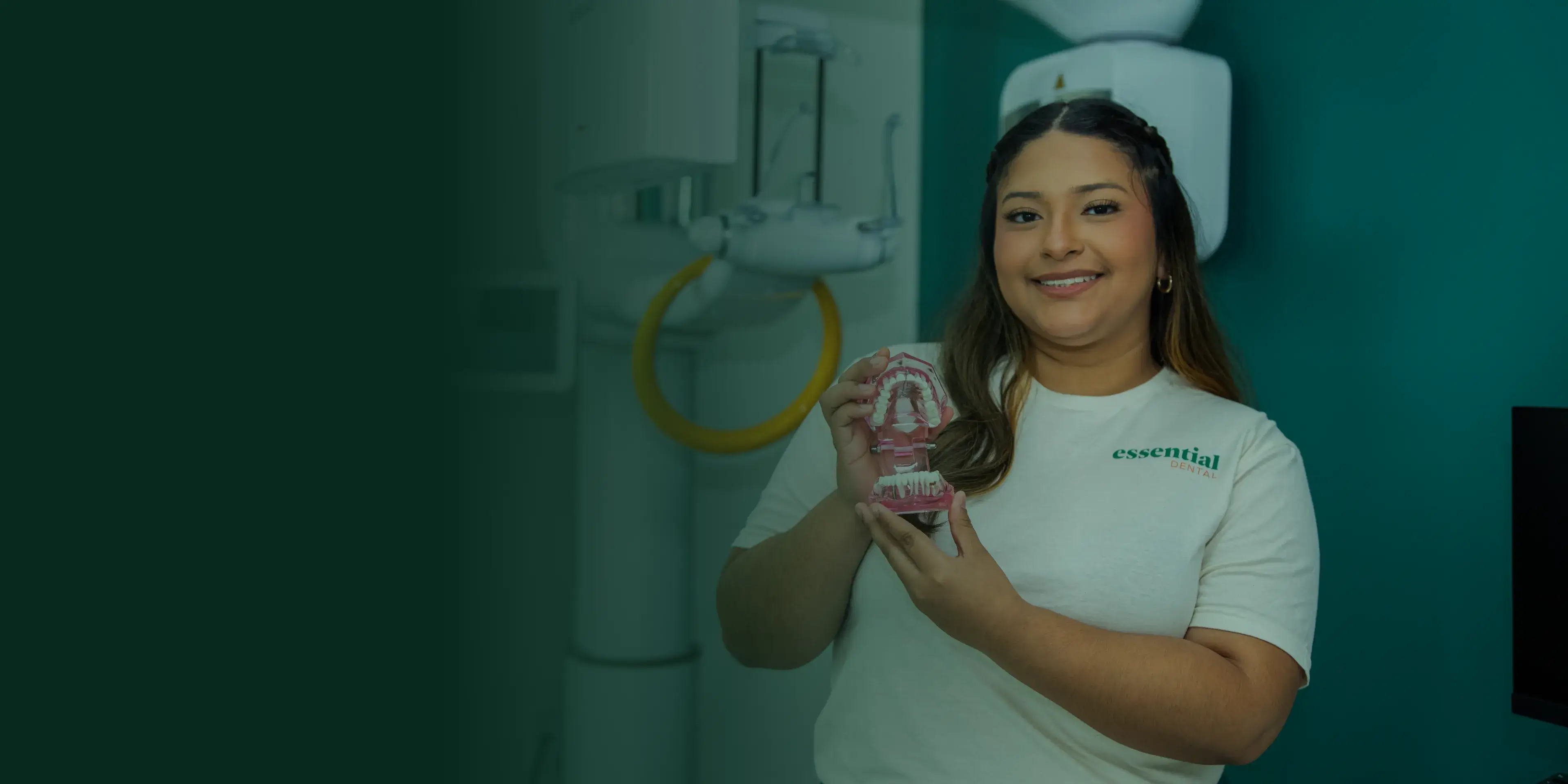 Smiling woman holding a dental model, standing in a dental office with equipment in the background.