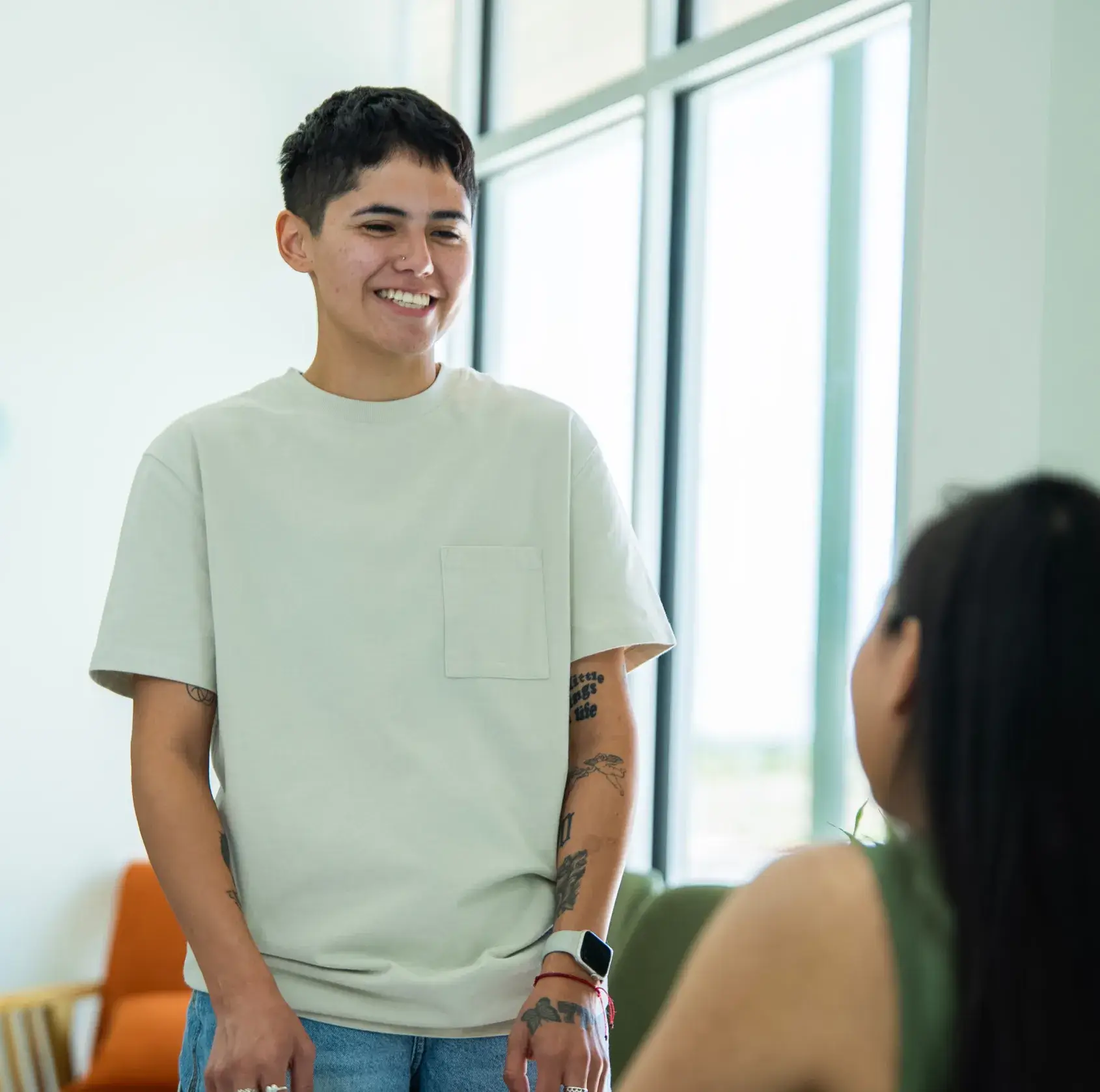 A person smiling while standing and talking to a seated person inside a bright room.