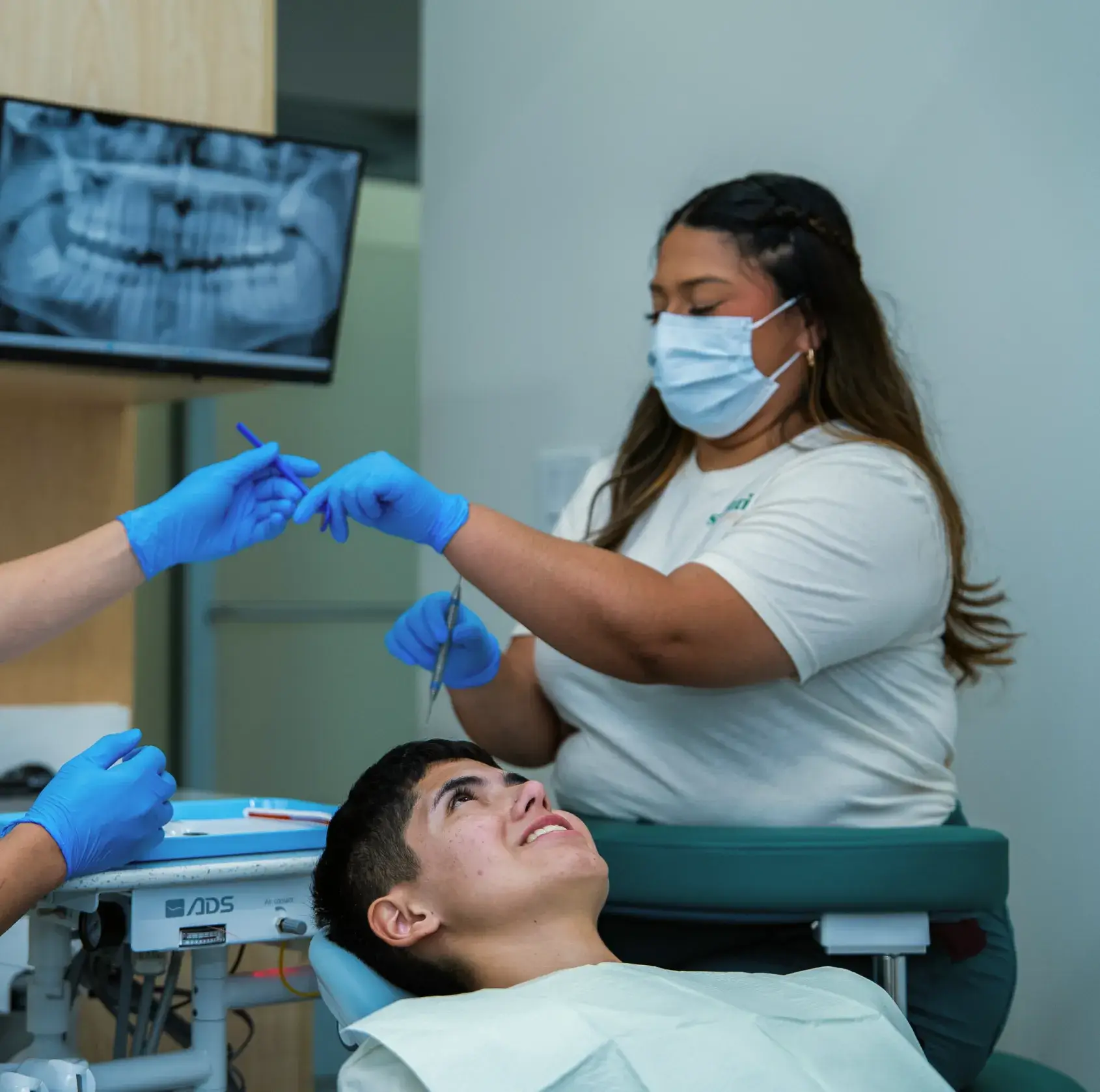 Patient sits in dental chair as dental assistant hands an instrument to another person.