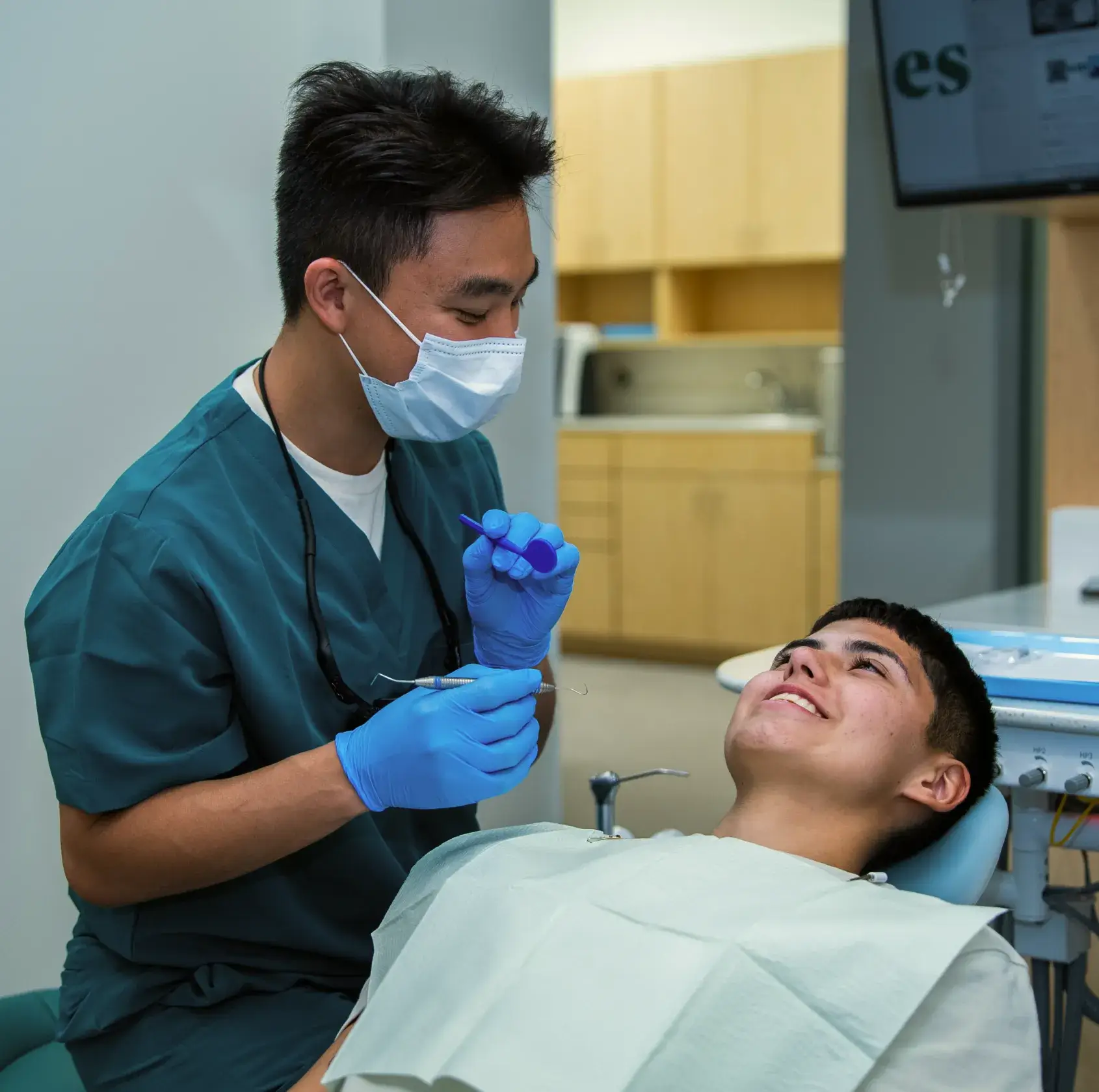 Dentist wearing a mask and gloves talks to a smiling patient lying in the dental chair.