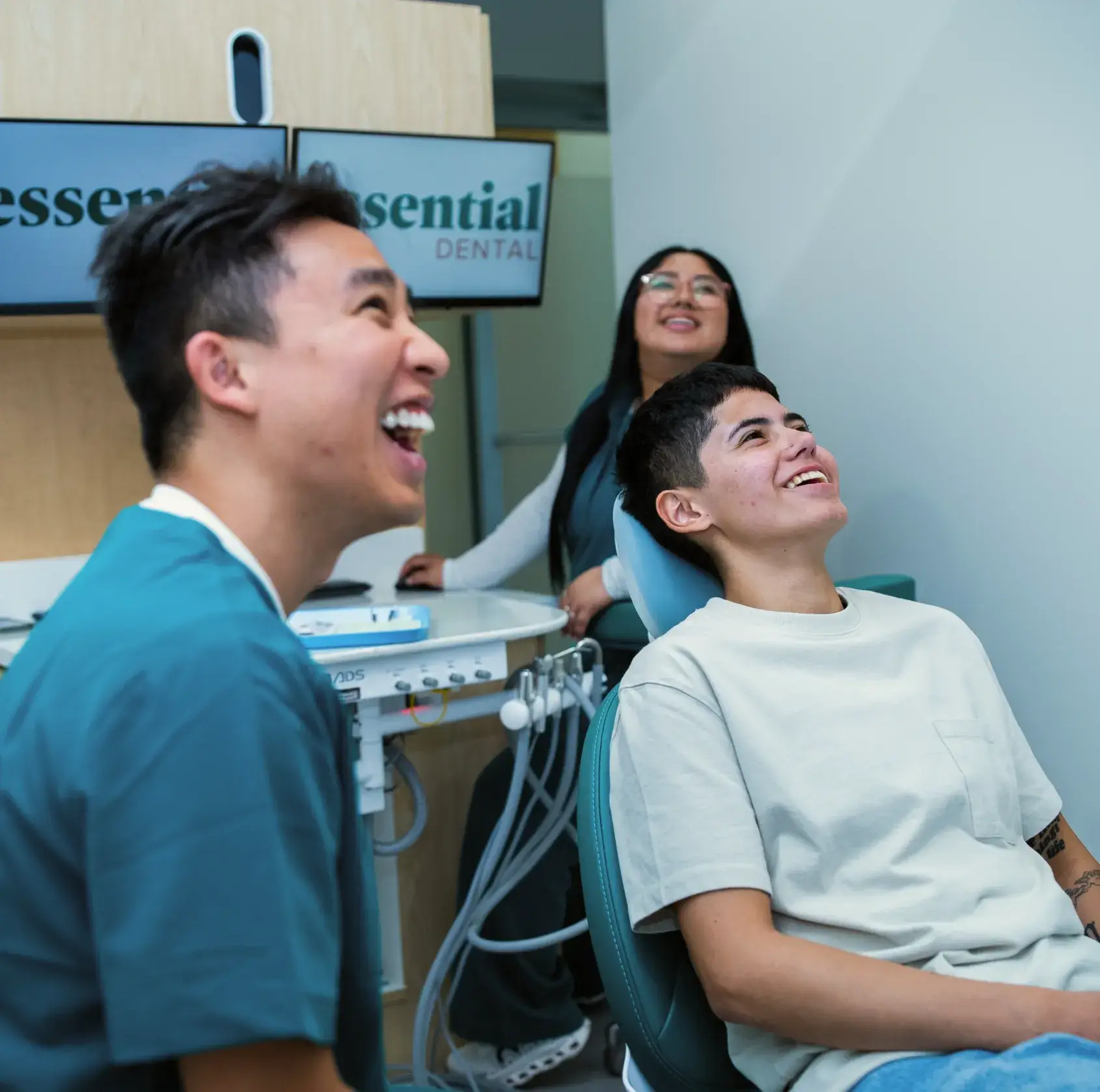 Two people in a dental office are laughing while one sits in the dental chair.