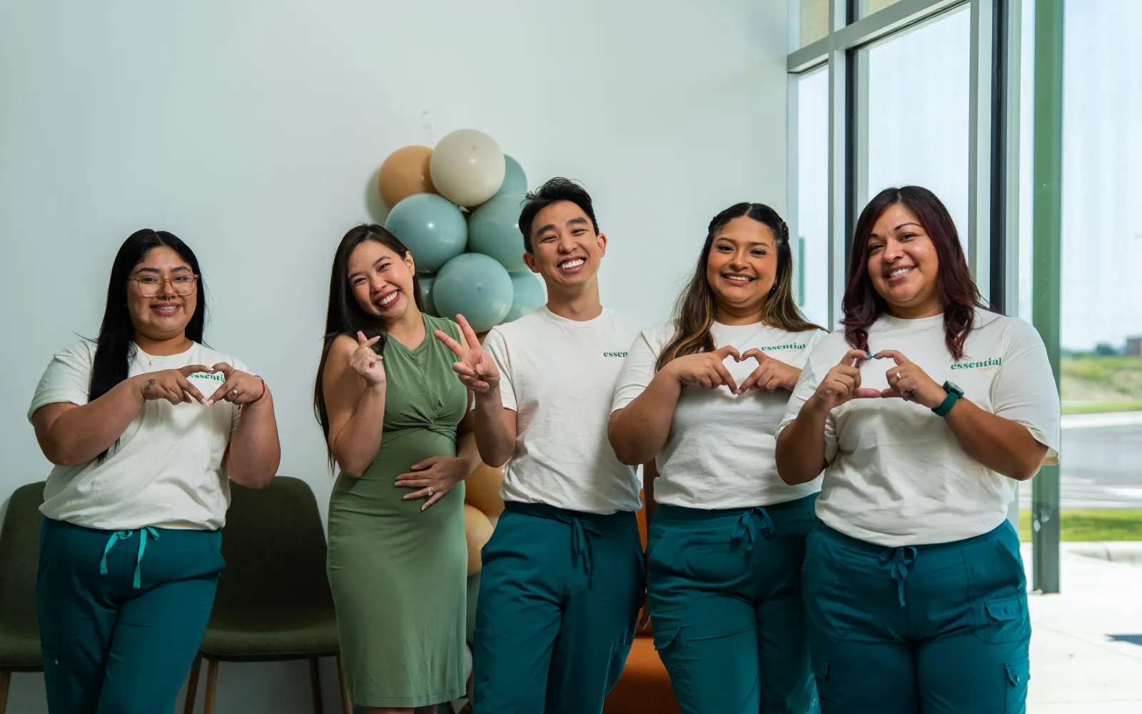 Five people stand smiling, making heart and peace signs with their hands in a bright room.
