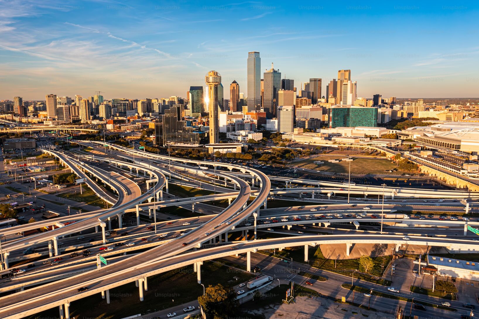 Aerial view of downtown Dallas, Texas, with a complex network of elevated highways in the foreground and the city skyline illuminated by the warm evening light.