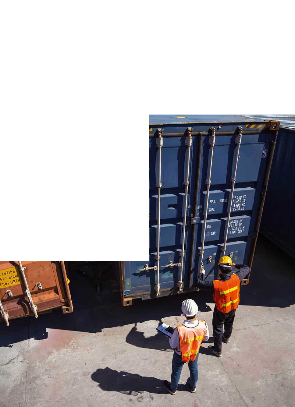Dwóch pracowników w kamizelkach odblaskowych i kaskach ocenia ładunek przy niebieskim kontenerze transportowym na placu.