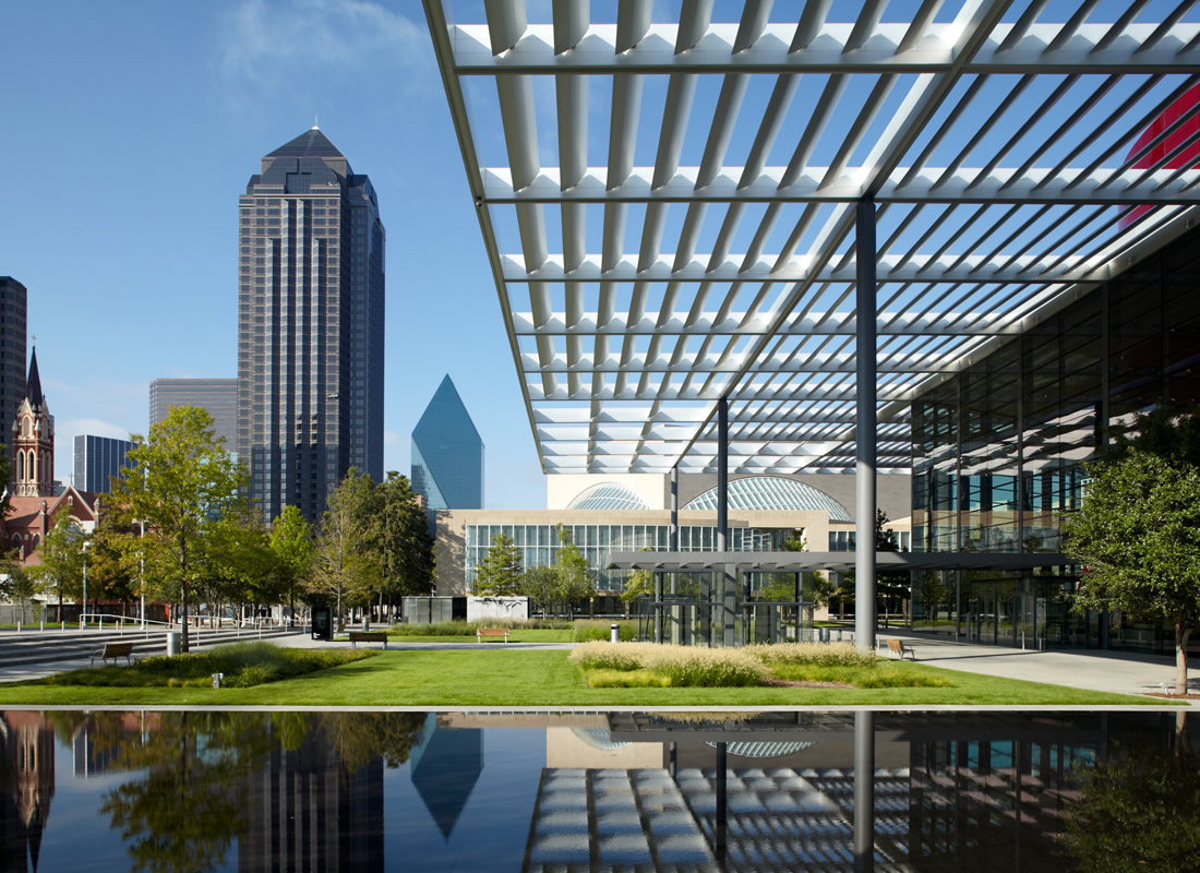 Modern urban park with green lawn and reflective pool in front of tall skyscrapers and a glass building with a grid-patterned overhang on a clear sunny day.