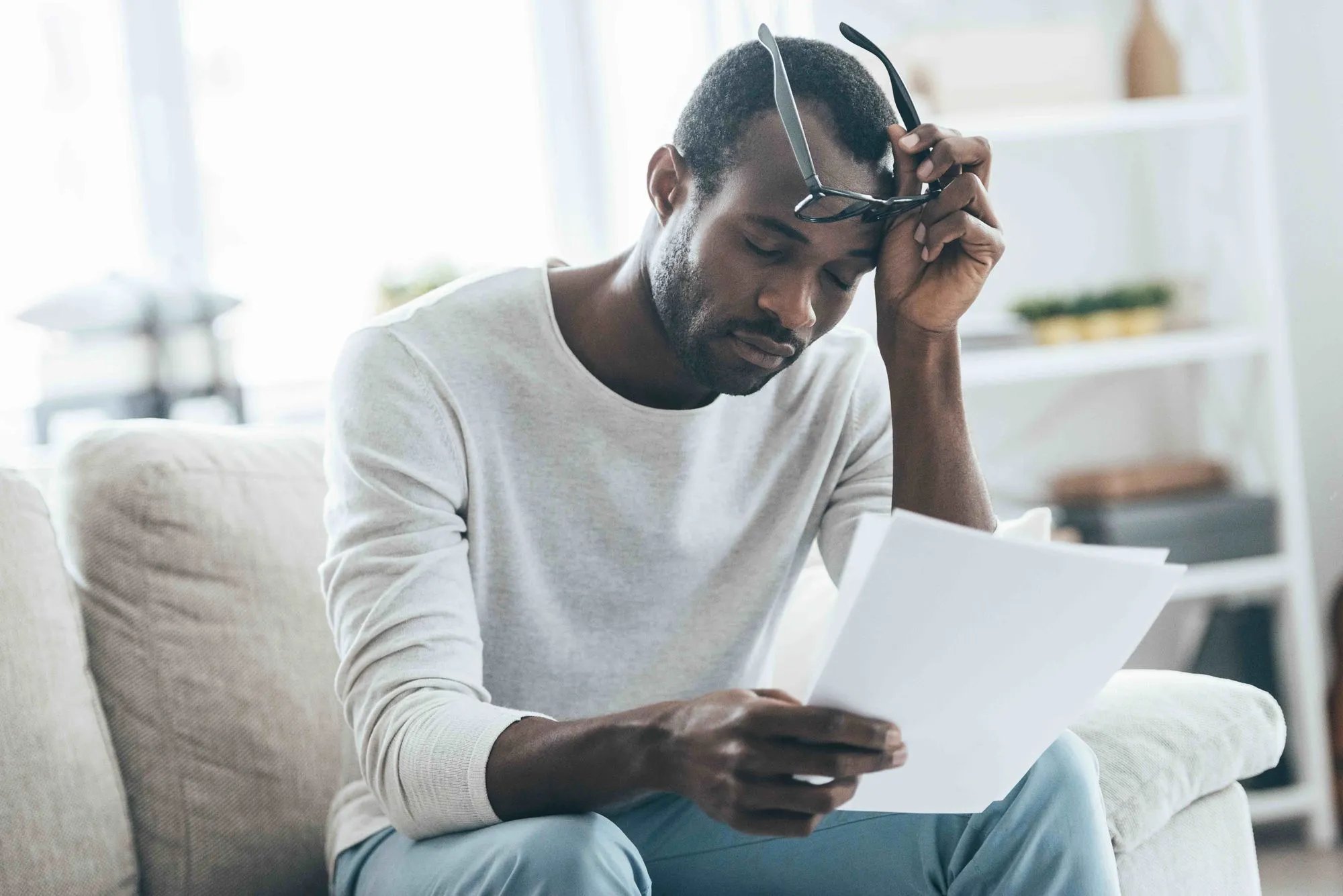 man sitting on a couch looking frustrated while reading paperwork