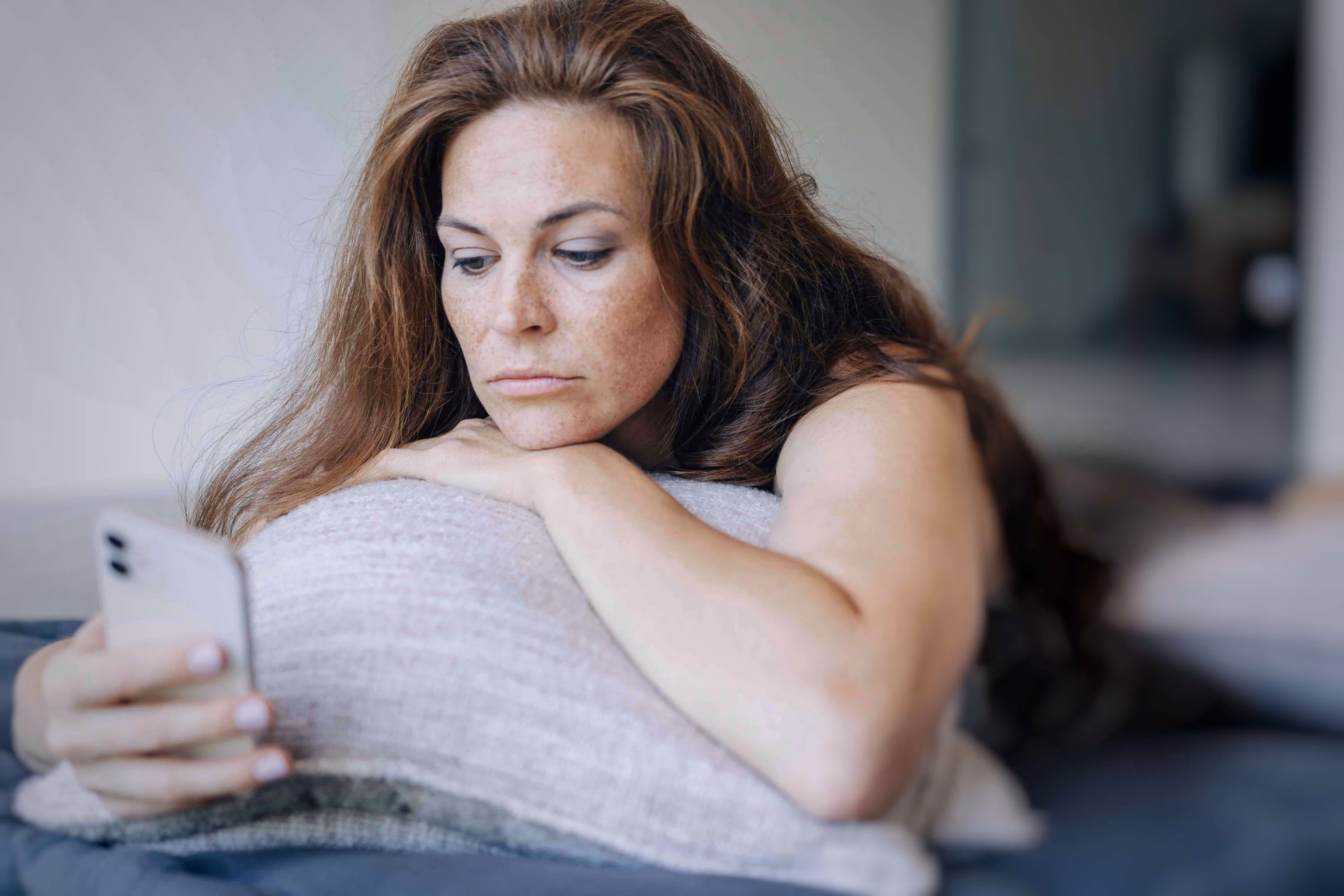 women laying on a pillow scrolling on her phone while looking sullen