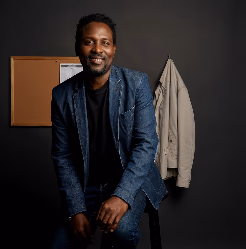 Man in a dark suit smiling in front of a black wall with a light-colored coat hanging beside him.