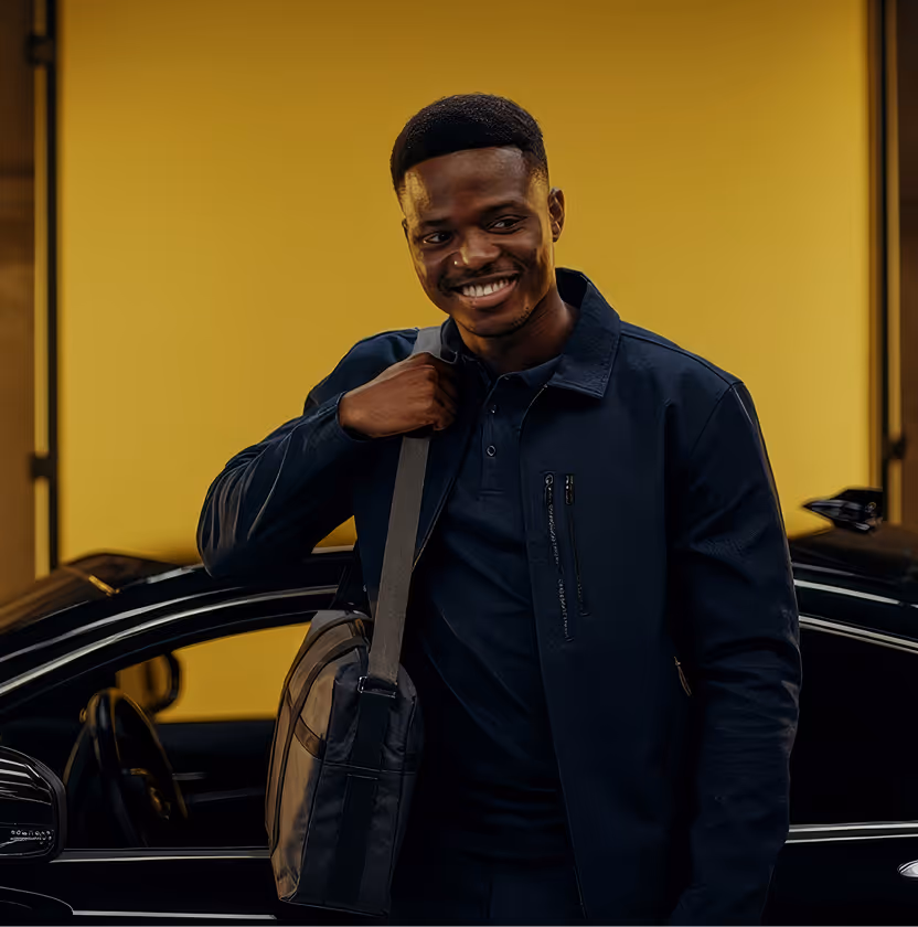 Man in a dark jacket smiling while stepping out of a black car in front of a yellow wall.
