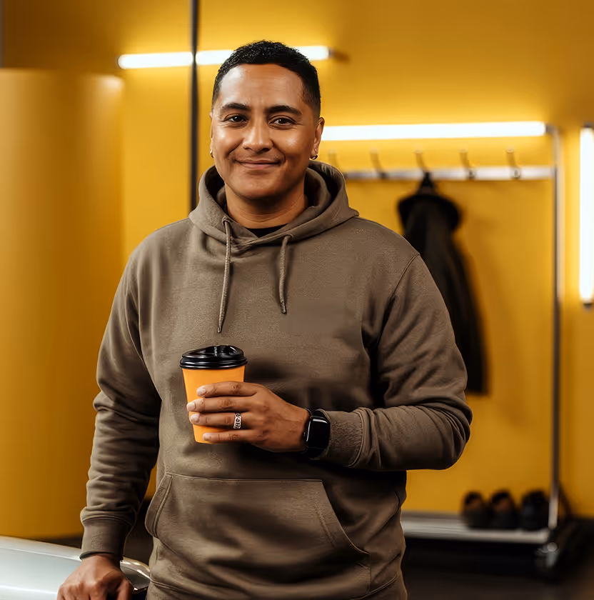 Man in a beige hoodie smiling and holding a cup in front of a yellow wall.