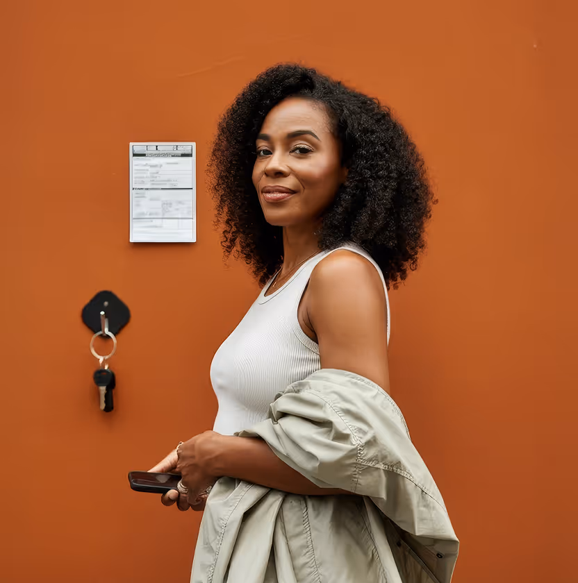 Smiling woman with curly hair, wearing a white top and holding a coat in front of an orange wall.
