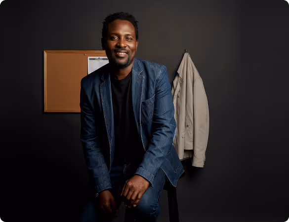 Man in a dark suit smiling in front of a black wall with a light-colored coat hanging beside him.