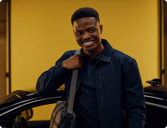 Man in a dark jacket smiling while stepping out of a black car in front of a yellow wall.