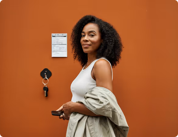 Smiling woman with curly hair, wearing a white top and holding a coat in front of an orange wall.