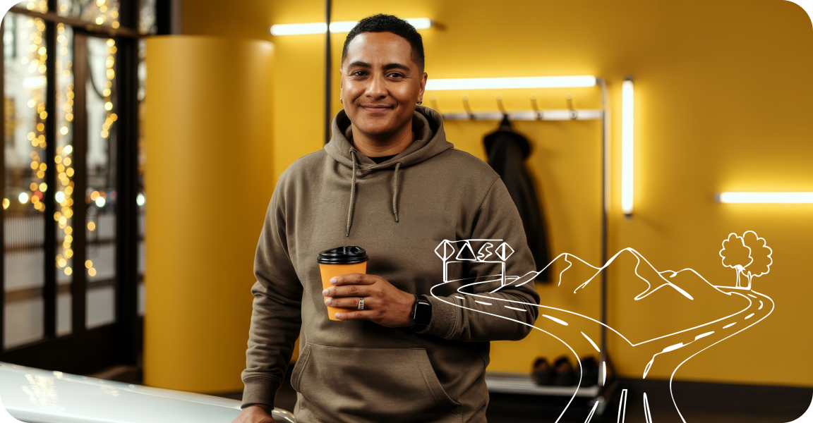 Man in a beige hoodie smiling and holding a cup in front of a yellow wall.