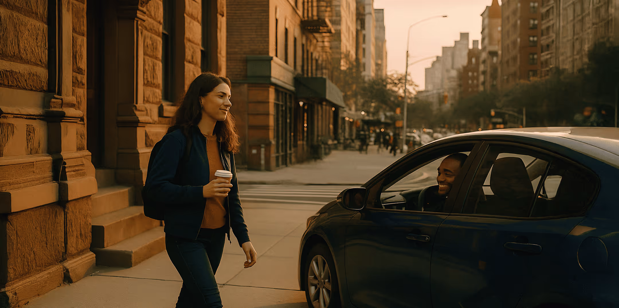 A young woman holding a coffee cup walks down a city sidewalk at sunset, smiling toward a man in a blue car parked nearby.