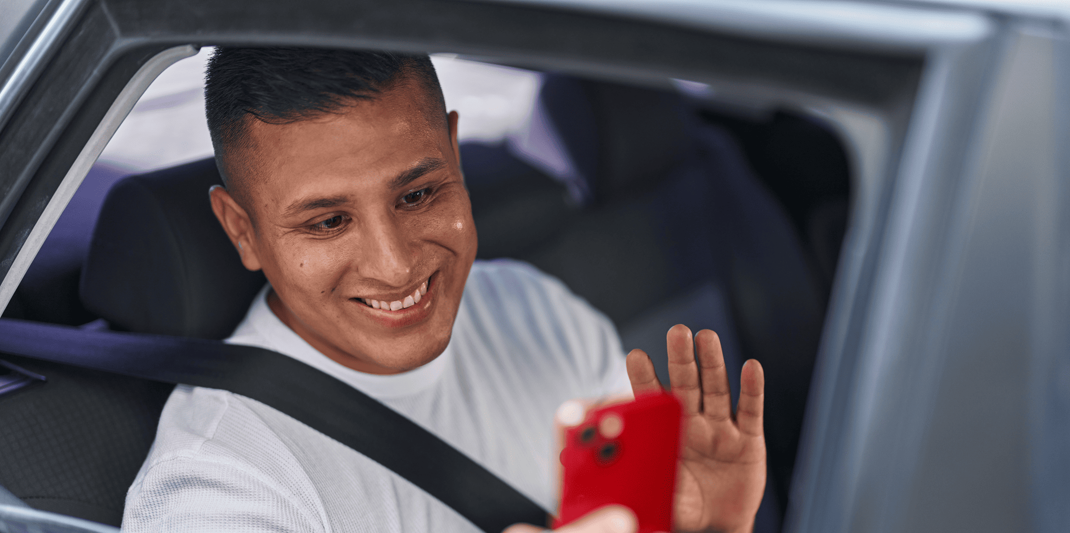 A man wearing a seatbelt and a white shirt smiles and waves while holding a red smartphone inside a car.