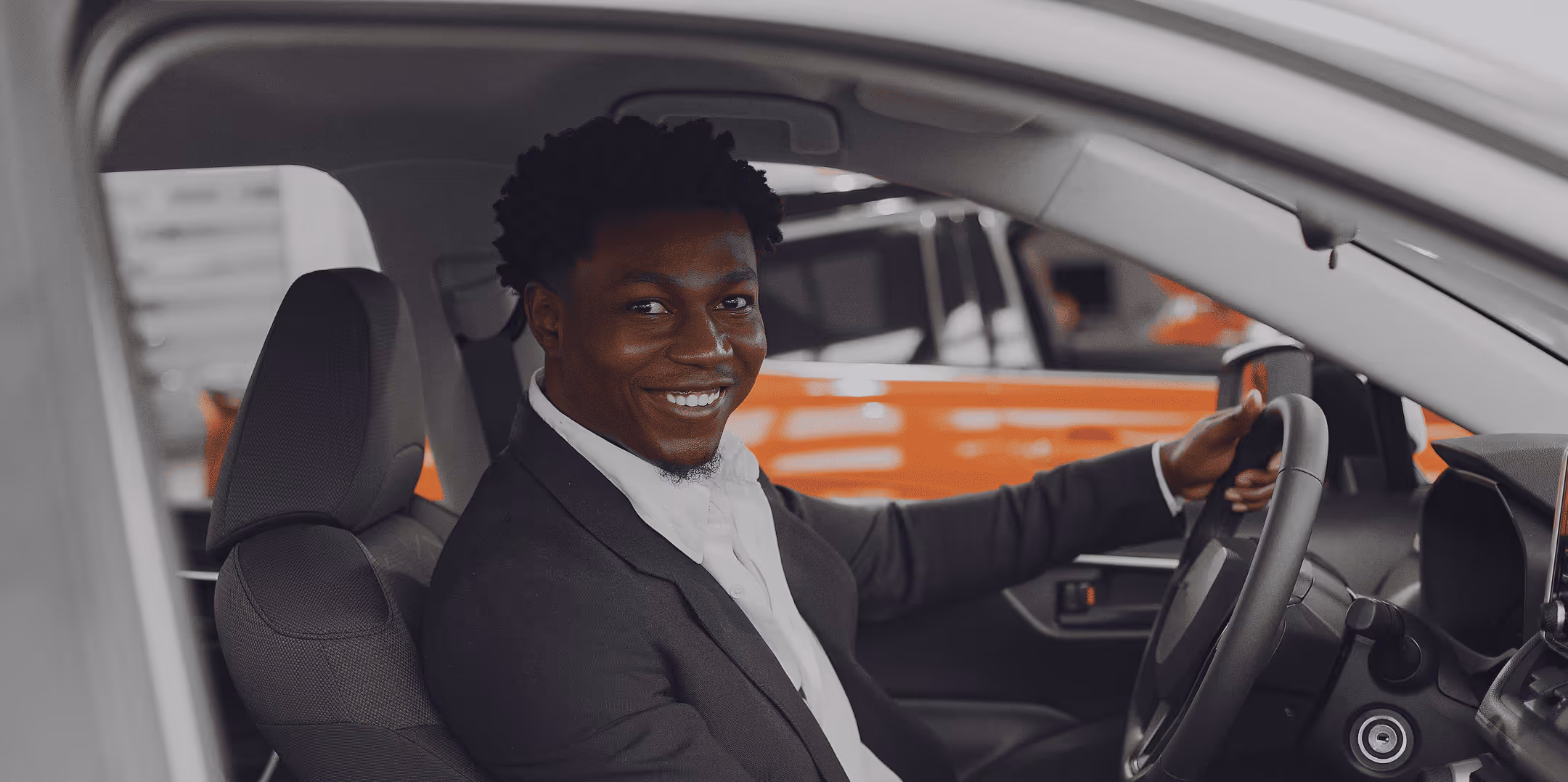 A man in a suit jacket sits in the driver’s seat of a car, smiling warmly at the camera.