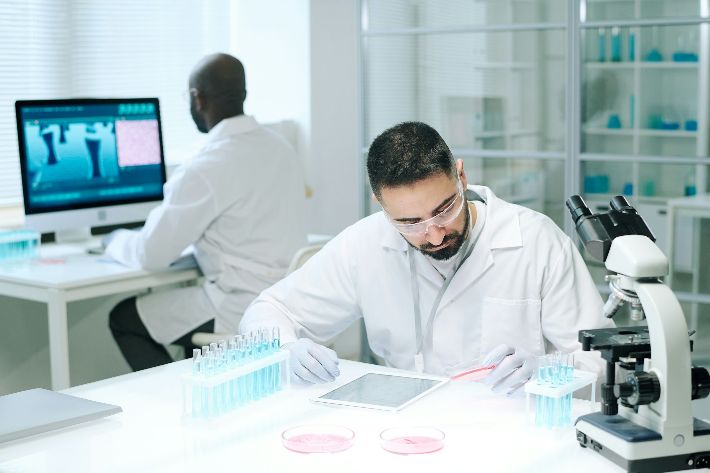 Young male scientist wearing protective goggles and gloves examining a petri dish in a laboratory with test tubes, a microscope, and a computer in the background.