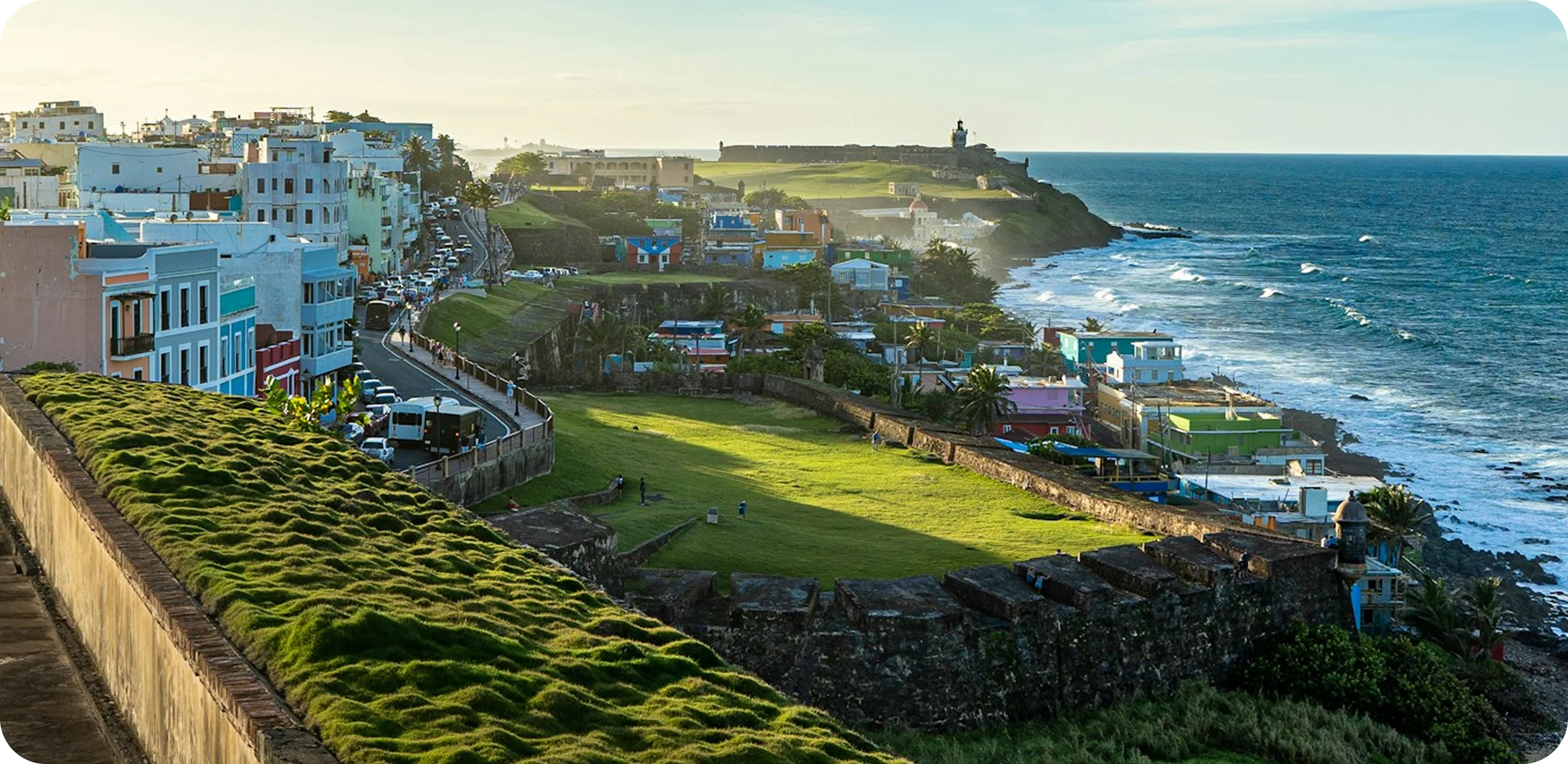 Scenic view of a historic coastal city with colorful buildings, green lawns on a fort wall, and the ocean waves crashing along the shore.