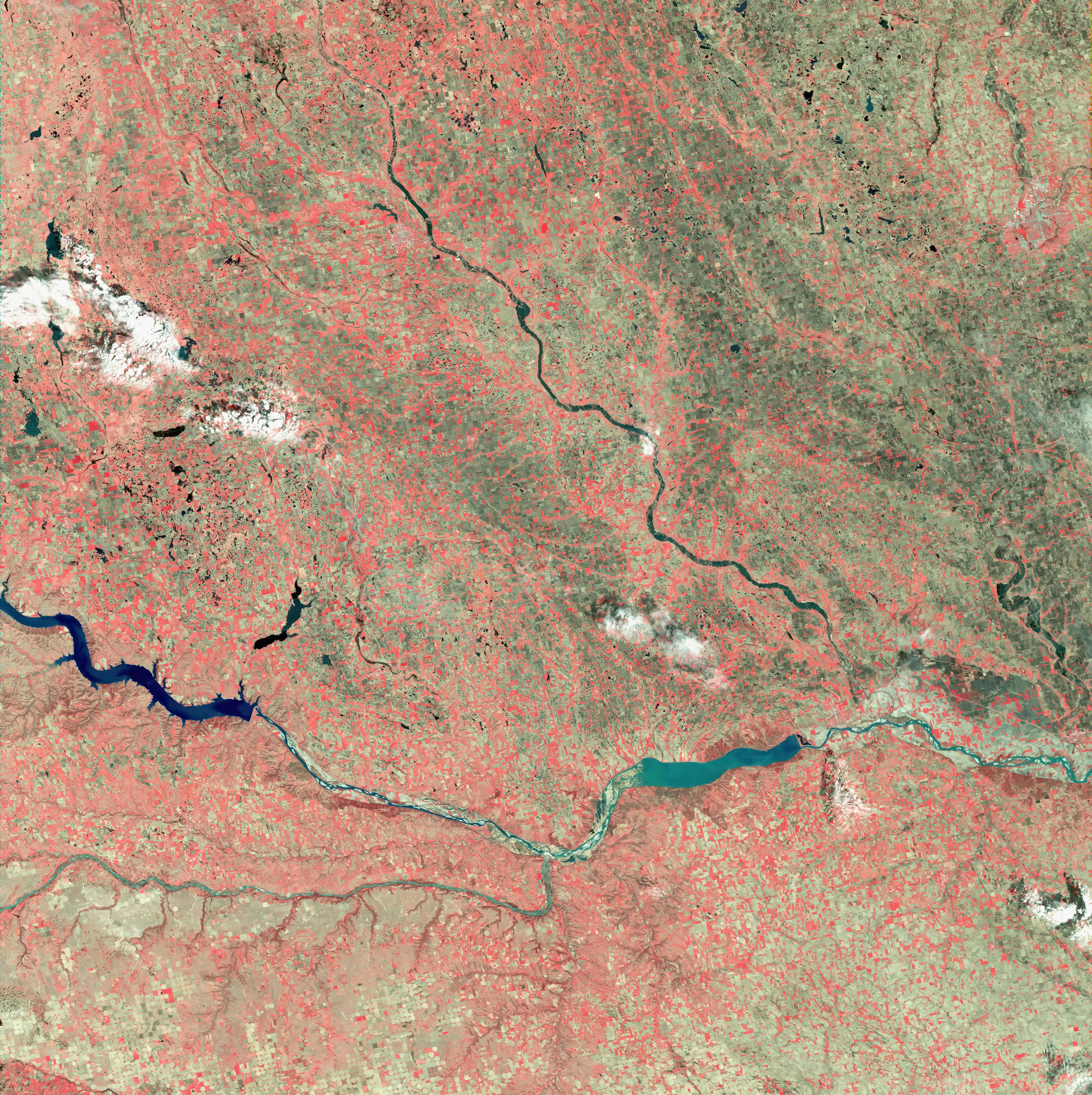 False-color satellite image showing a winding river cutting through a patchwork of agricultural fields, with red tones indicating vegetation. Scattered white clouds are visible across the landscape, and the terrain appears mostly flat with some hilly areas.