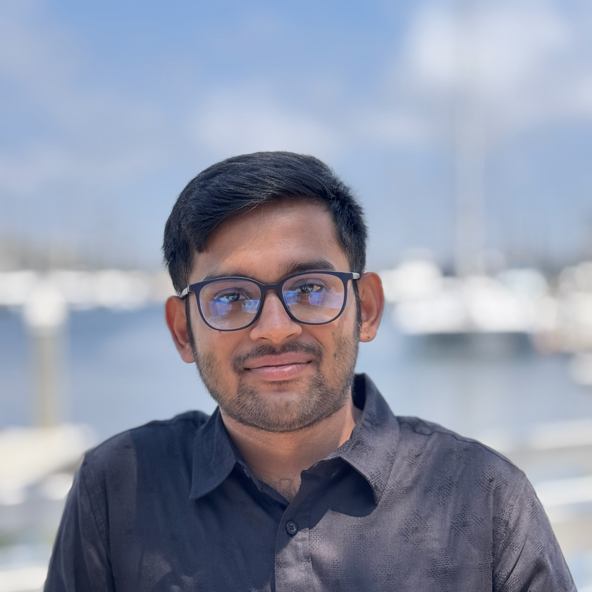 Jay Famiglietti, a man with short gray hair, a neatly trimmed beard, and glasses, smiling in a plaid blazer and collared shirt, standing outdoors with blurred architecture and greenery in the background.