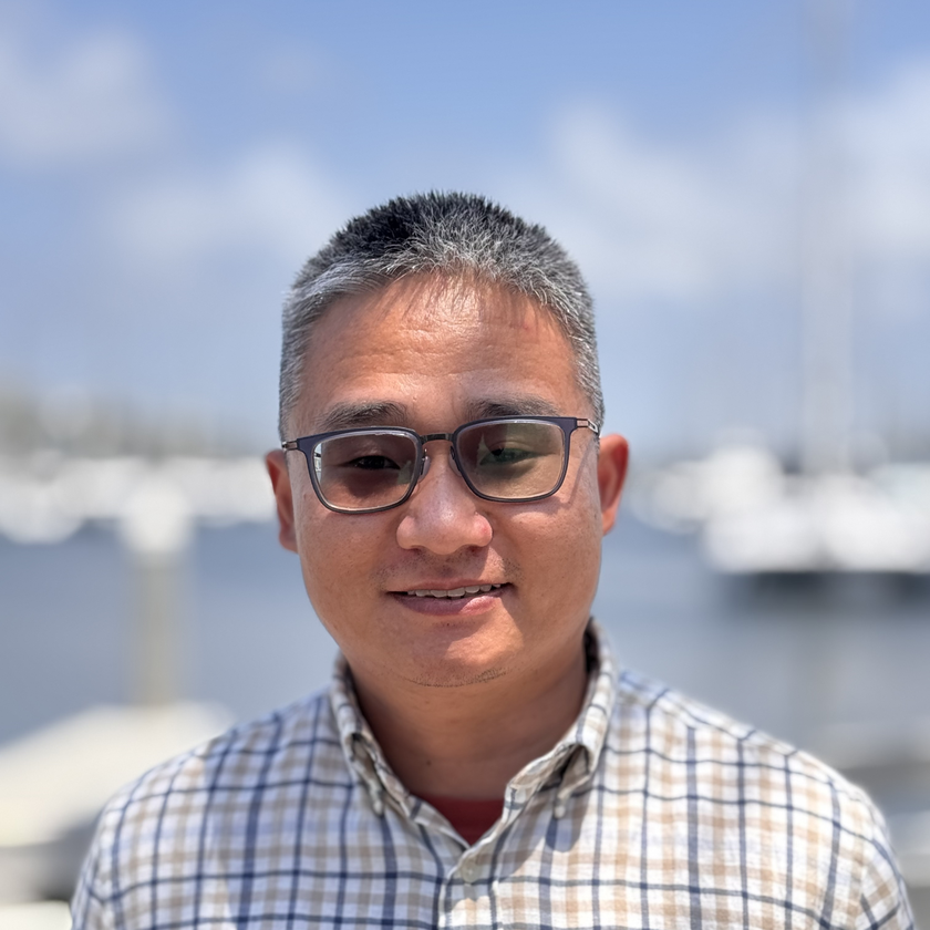 Jay Famiglietti, a man with short gray hair, a neatly trimmed beard, and glasses, smiling in a plaid blazer and collared shirt, standing outdoors with blurred architecture and greenery in the background.