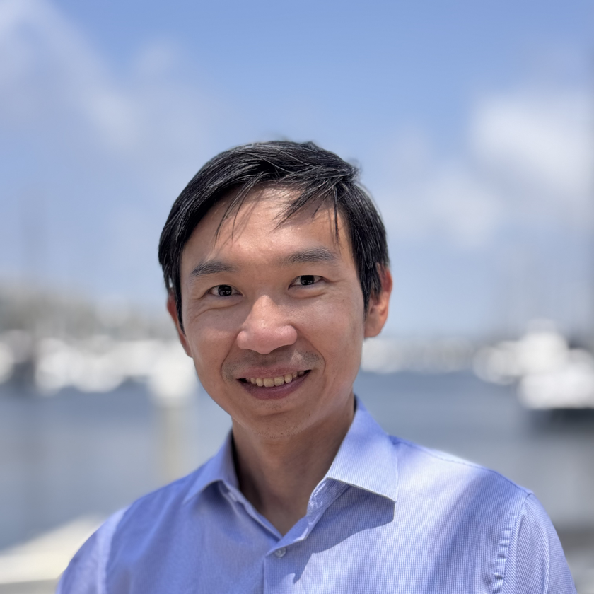 Jay Famiglietti, a man with short gray hair, a neatly trimmed beard, and glasses, smiling in a plaid blazer and collared shirt, standing outdoors with blurred architecture and greenery in the background.