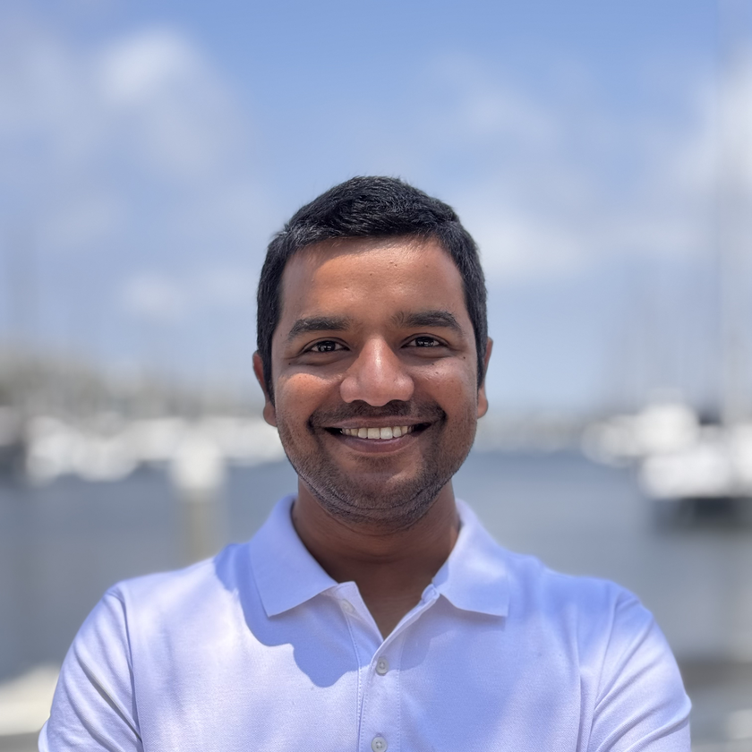 Jay Famiglietti, a man with short gray hair, a neatly trimmed beard, and glasses, smiling in a plaid blazer and collared shirt, standing outdoors with blurred architecture and greenery in the background.