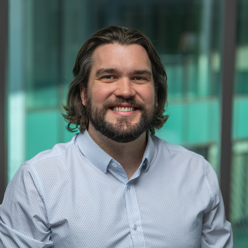 Jay Famiglietti, a man with short gray hair, a neatly trimmed beard, and glasses, smiling in a plaid blazer and collared shirt, standing outdoors with blurred architecture and greenery in the background.