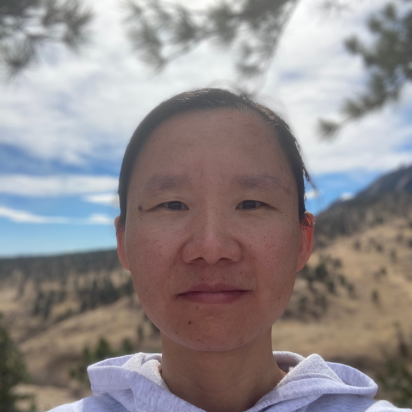 Jay Famiglietti, a man with short gray hair, a neatly trimmed beard, and glasses, smiling in a plaid blazer and collared shirt, standing outdoors with blurred architecture and greenery in the background.