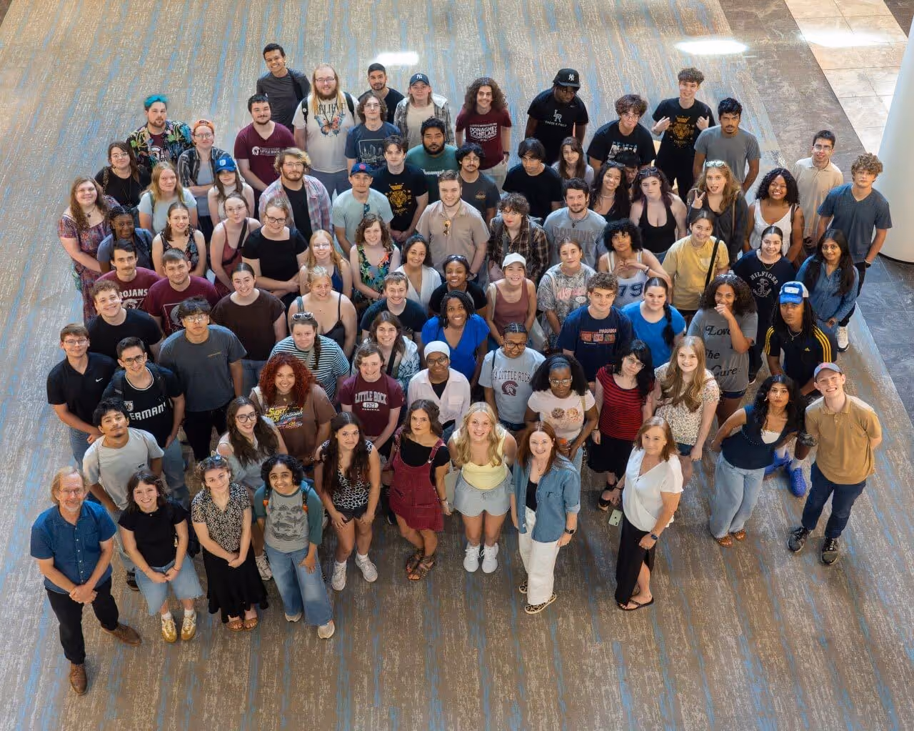 Group of around 70 Donaghey Scholars gathered indoors, standing close together and looking up at the camera with smiles and casual attire.
