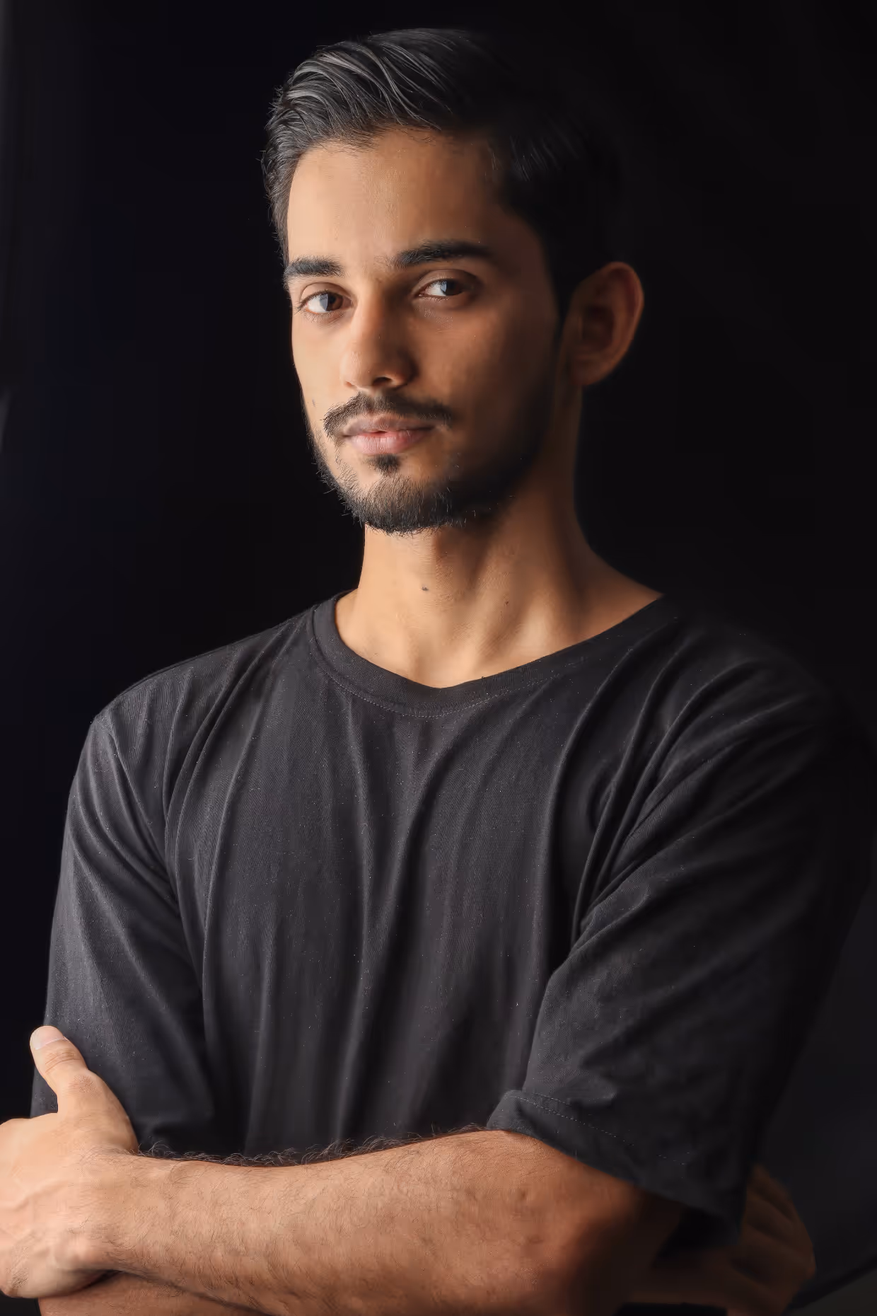 Young man with short dark hair and beard, wearing a black t-shirt, standing with arms crossed against a black background.