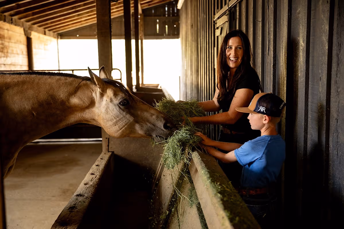 A woman and a boy feeding Anderson Hay to a horse in a horse barn