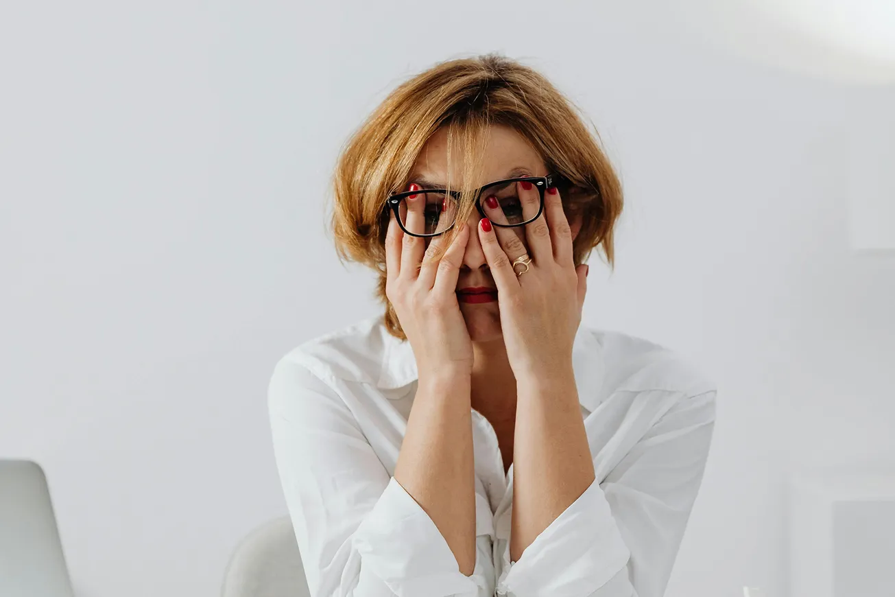 A woman wearing glasses sitting at a desk with her hands on her face.