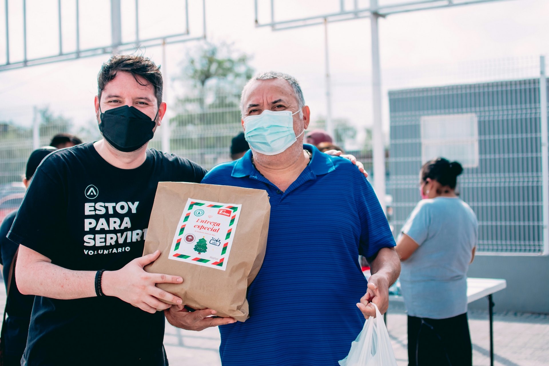 Two men wearing masks stand outdoors, smiling and holding a brown paper bag labeled "Entrega especial." The mood is warm and communal.