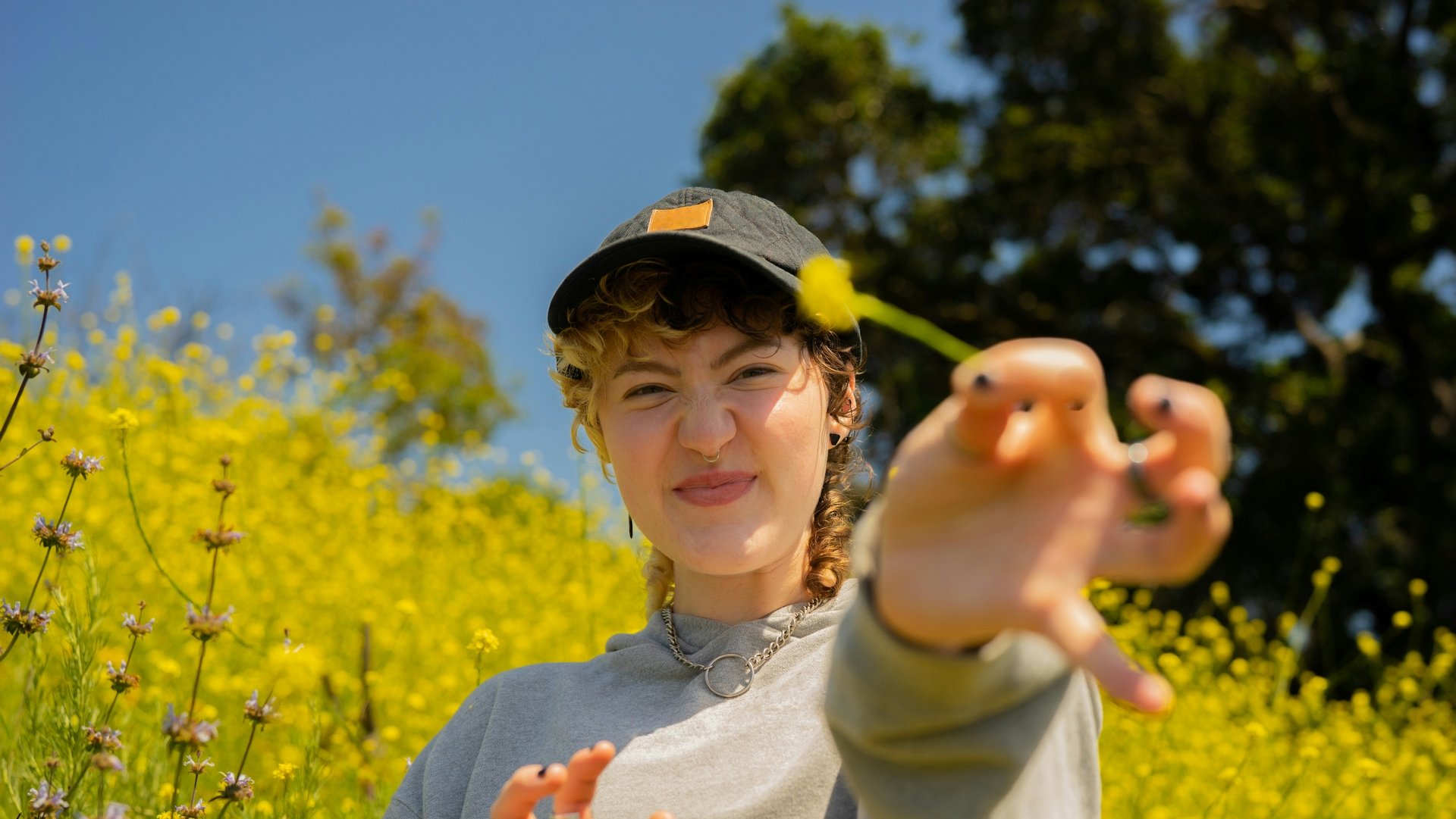 Young person in gray sweatshirt and cap smiling playfully, reaching toward the camera. They're surrounded by a vibrant field of yellow wildflowers under a clear blue sky.