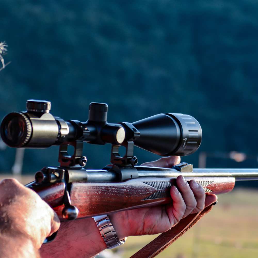 A man stands holding a rifle, looking serious and focused, with a neutral background.