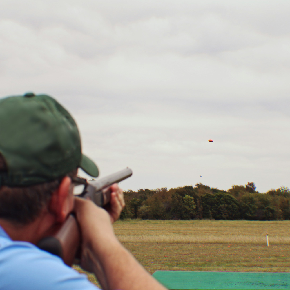 A man wearing ear protection aiming a shotgun at a clay pigeon in a shooting range setting.