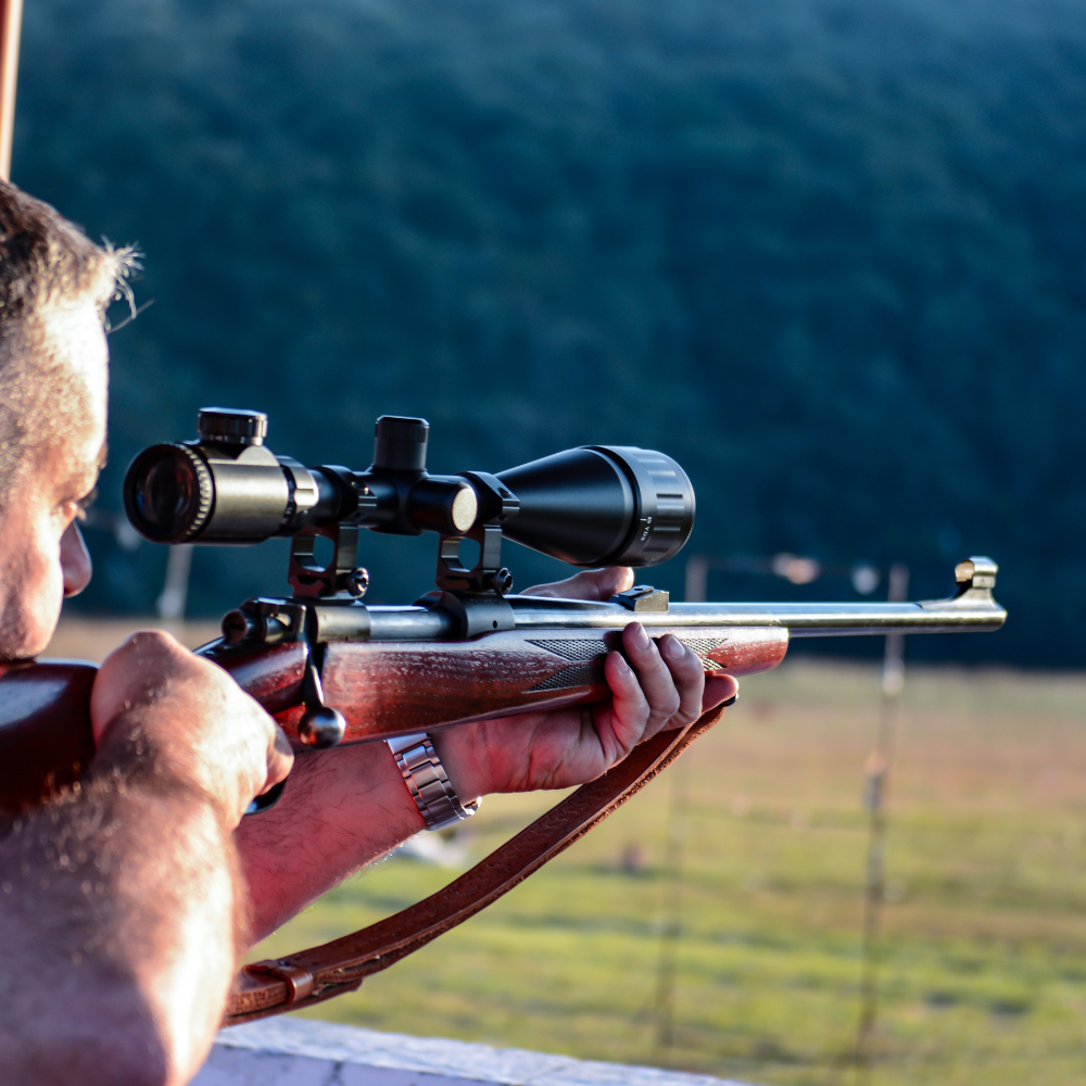 A man stands holding a rifle, looking serious and focused, with a neutral background.