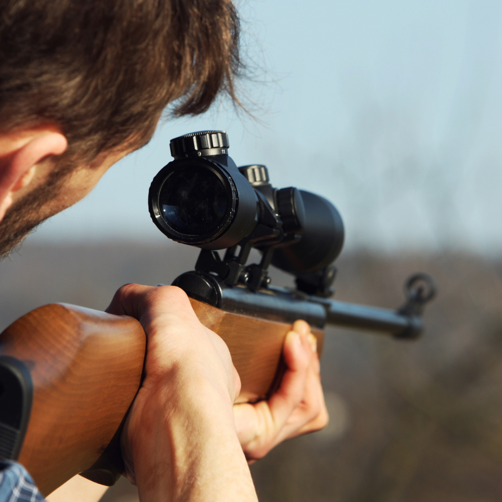 A man stands holding a rifle, looking serious and focused, with a neutral background.