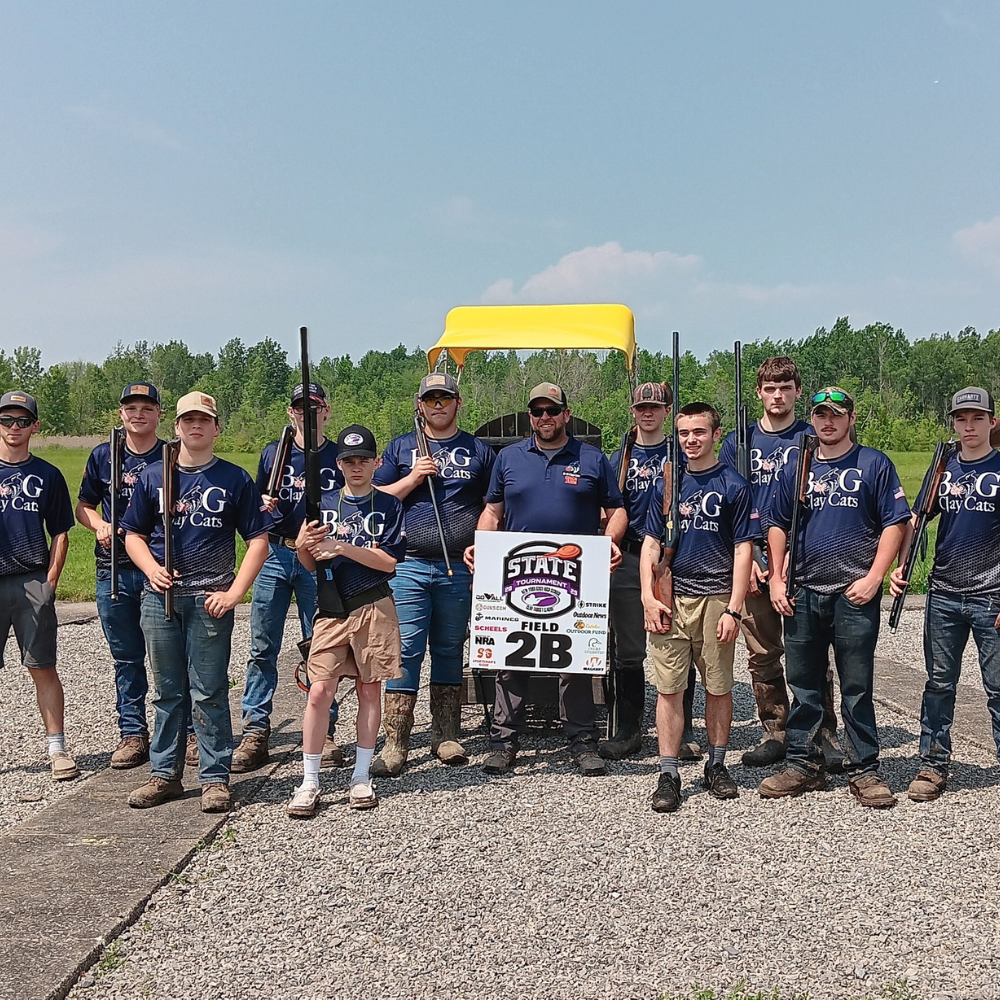 A group of BG Clay Cat members in blue shirts and jeans stand outdoors holding shotguns, posing with a “State Field 2B” sign and a yellow golf cart behind them. Trees and sky are visible in the background.