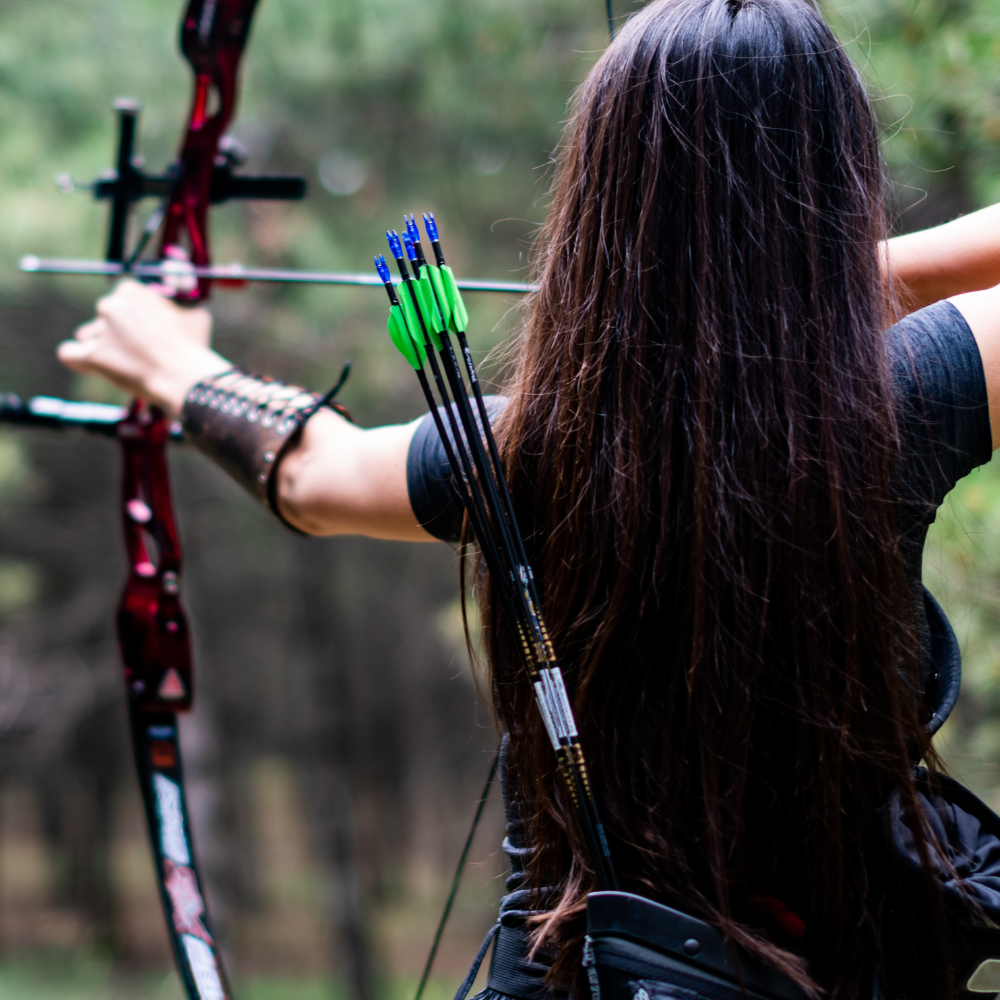 A woman with long hair is aiming a bow in a forest, focused and poised. Bright green arrows are in a quiver. The scene is calm and serene.