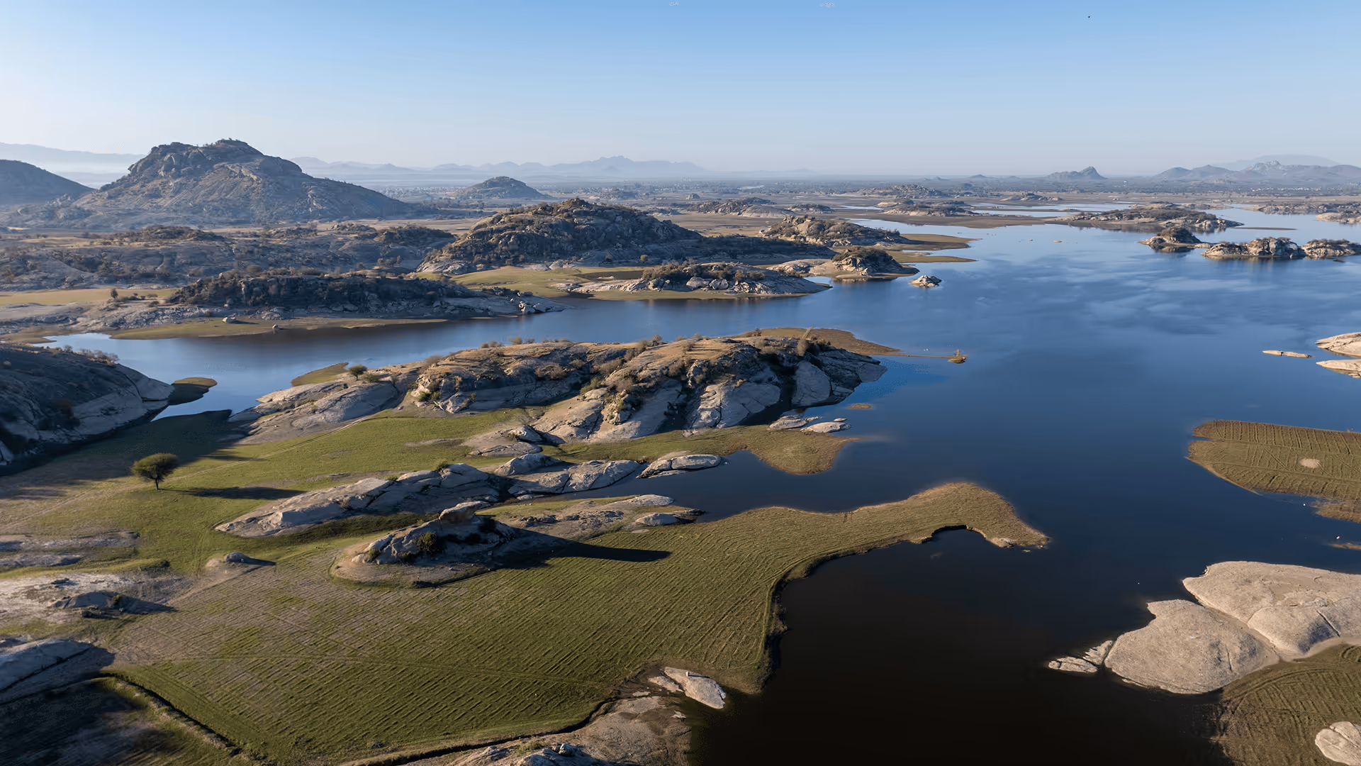Aerial view of a serene lake surrounded by rocky hills and patches of green grass under a clear blue sky.