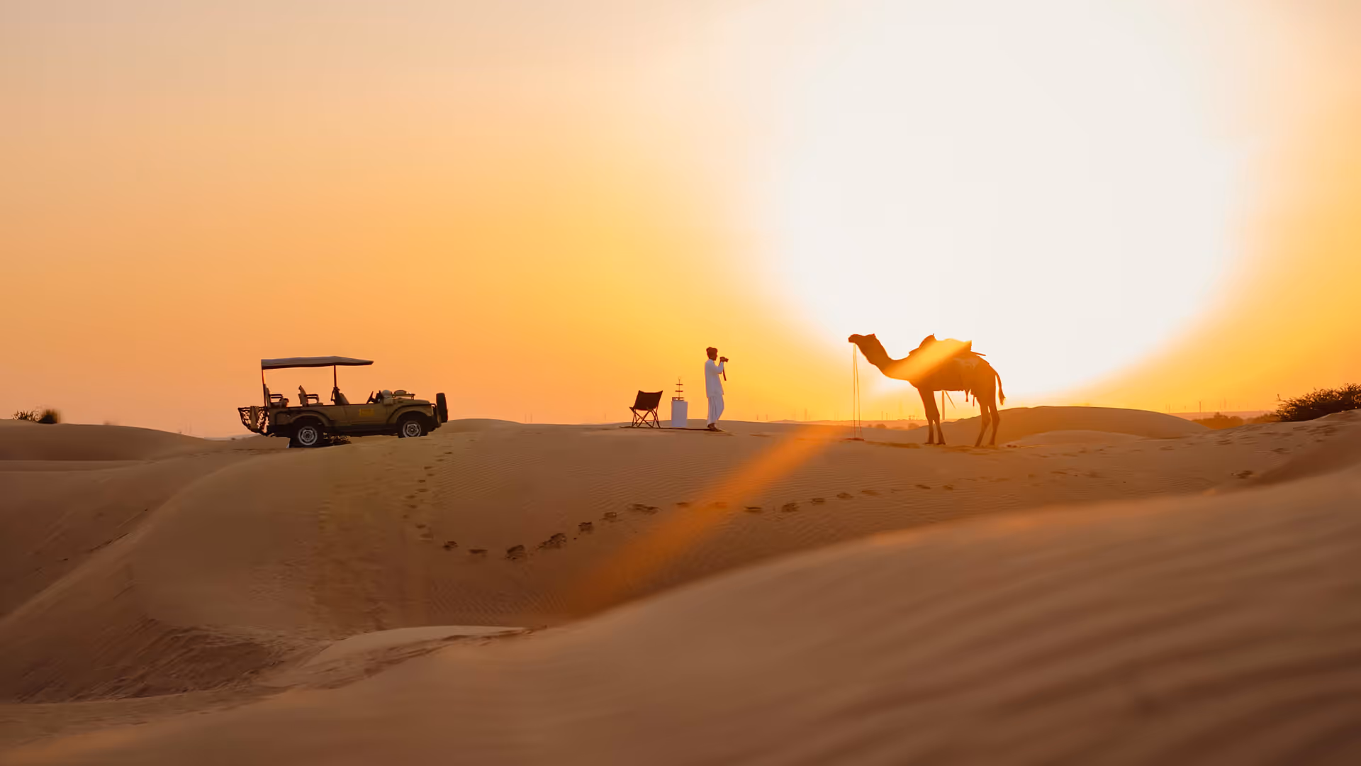 Sunset desert scene with a person in white taking a photo, a camel, a safari vehicle, and sand dunes.