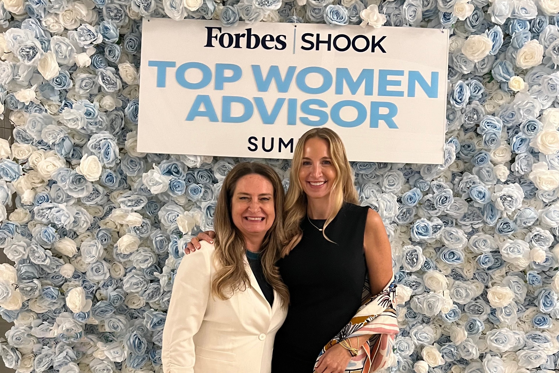 Lara Rhame and Kirsten Pickens in front of the Forbes Shook Top Women Advisor Summit sign and wall of roses.